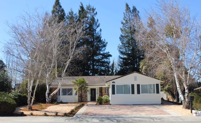 Single-story ranch-style house with a driveway, landscaped front yard and trees under a clear blue sky.