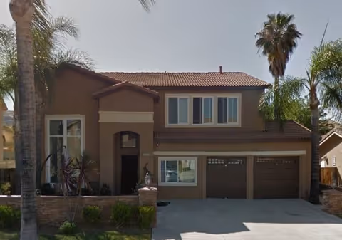 Two-story brown stucco house with a two-car garage and palm trees.