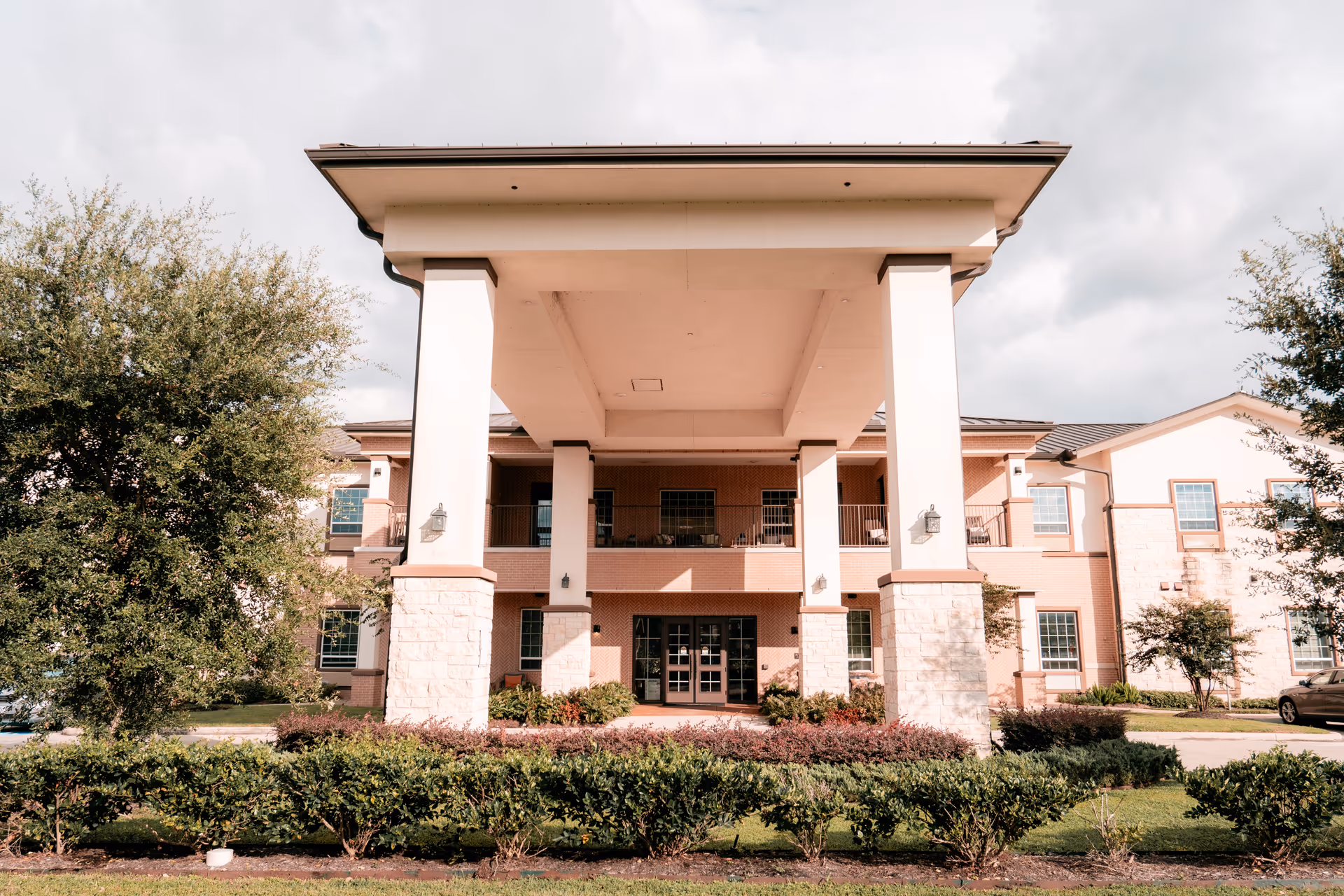 Front exterior of a senior living building featuring a large covered entry portico with columns, balconies, and surrounding landscaping.