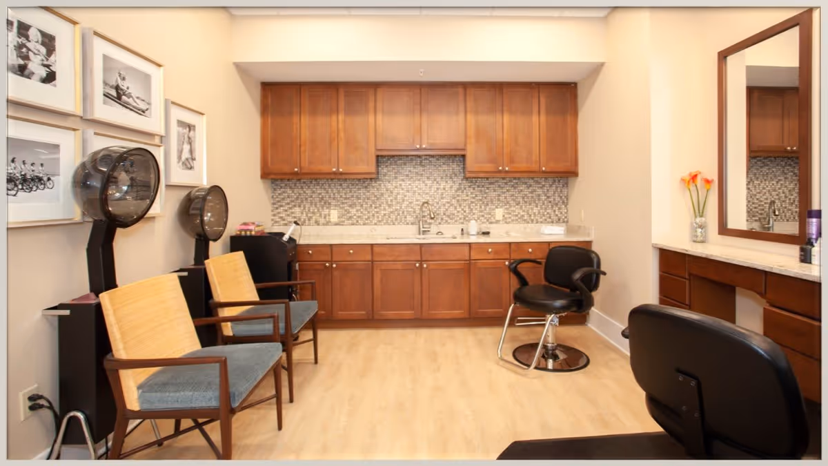 Interior view of a hair salon area in a senior living facility with wooden cabinets, a countertop with a sink, two hair drying stations with chairs, and a styling chair in front of a large mirror. The walls are decorated with black and white framed photographs.