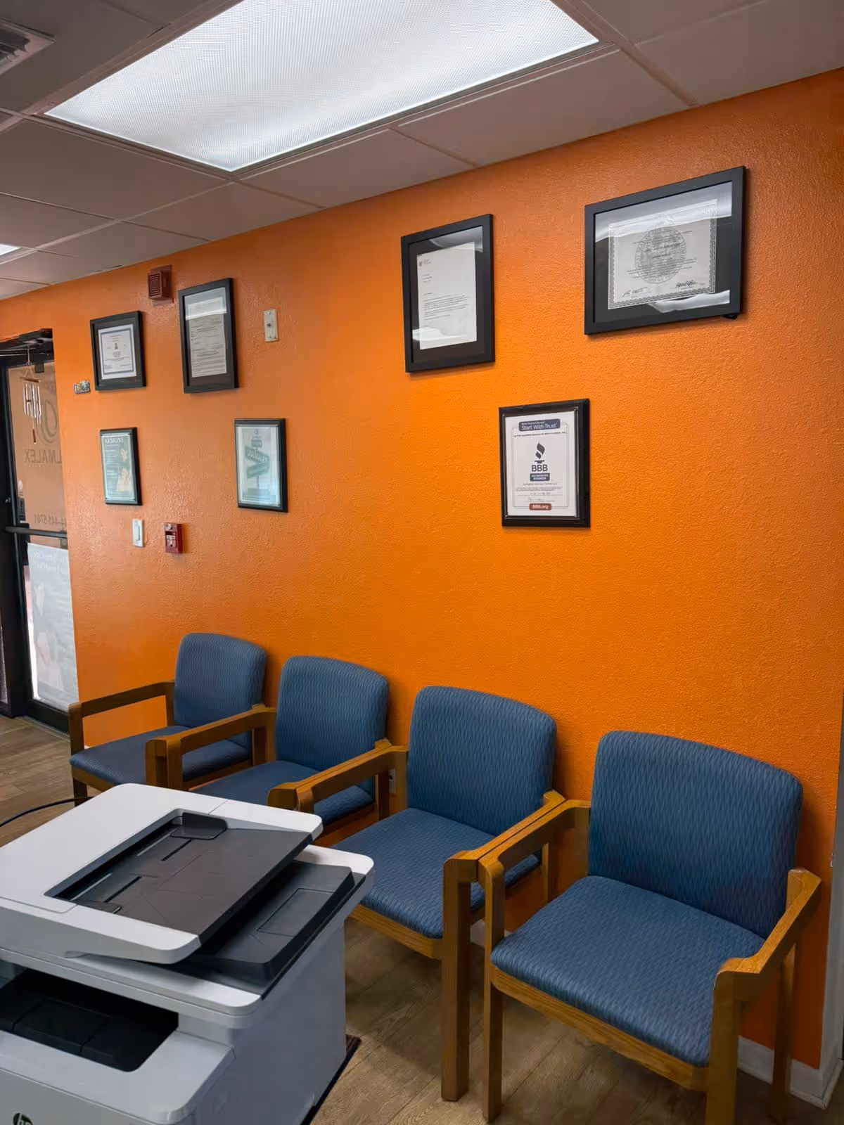 Orange-walled waiting area with blue chairs, framed certificates, and a printer.