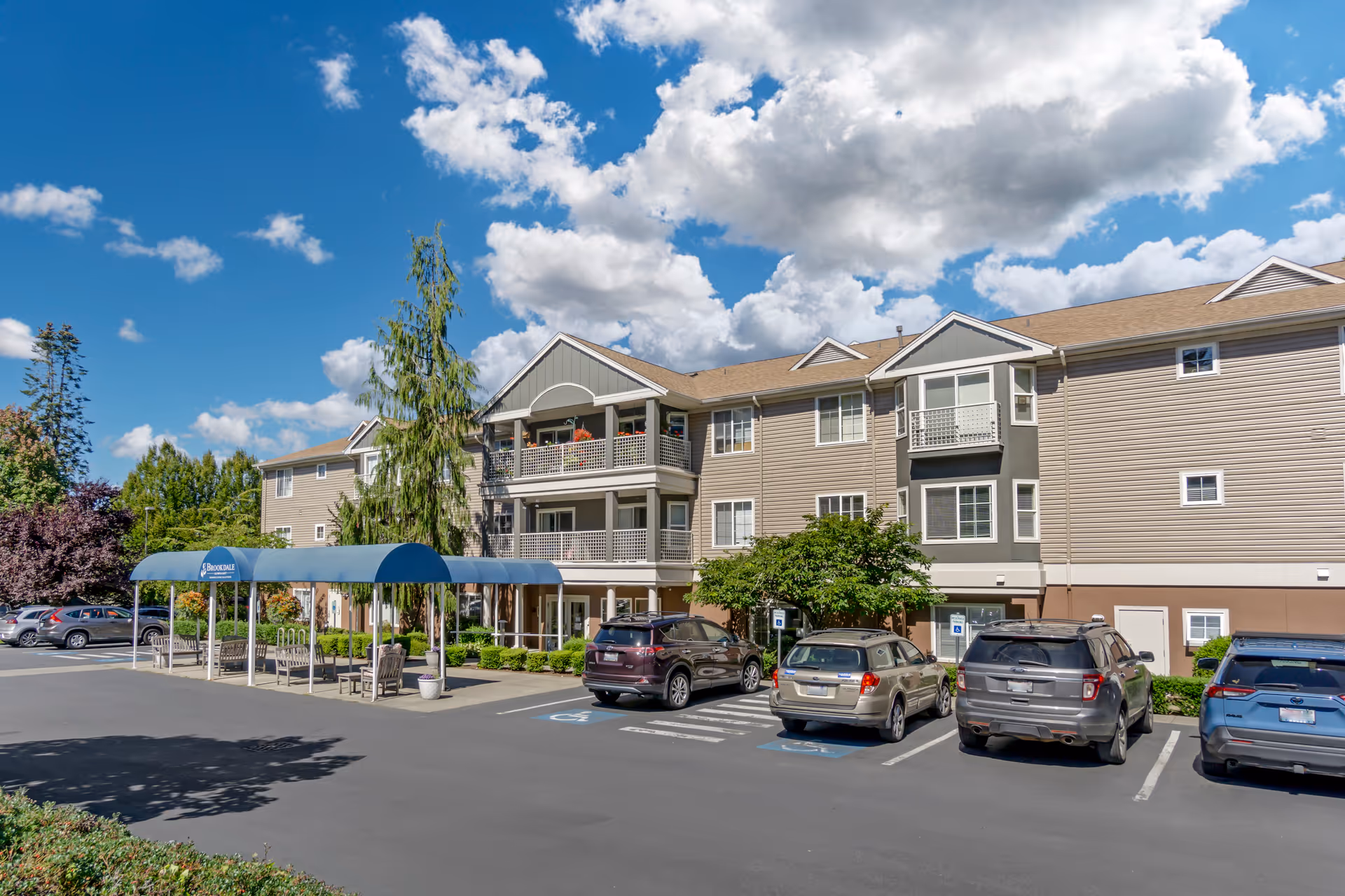 Three-story senior living building with a covered entrance canopy, parked cars in the lot, and a blue sky with clouds.