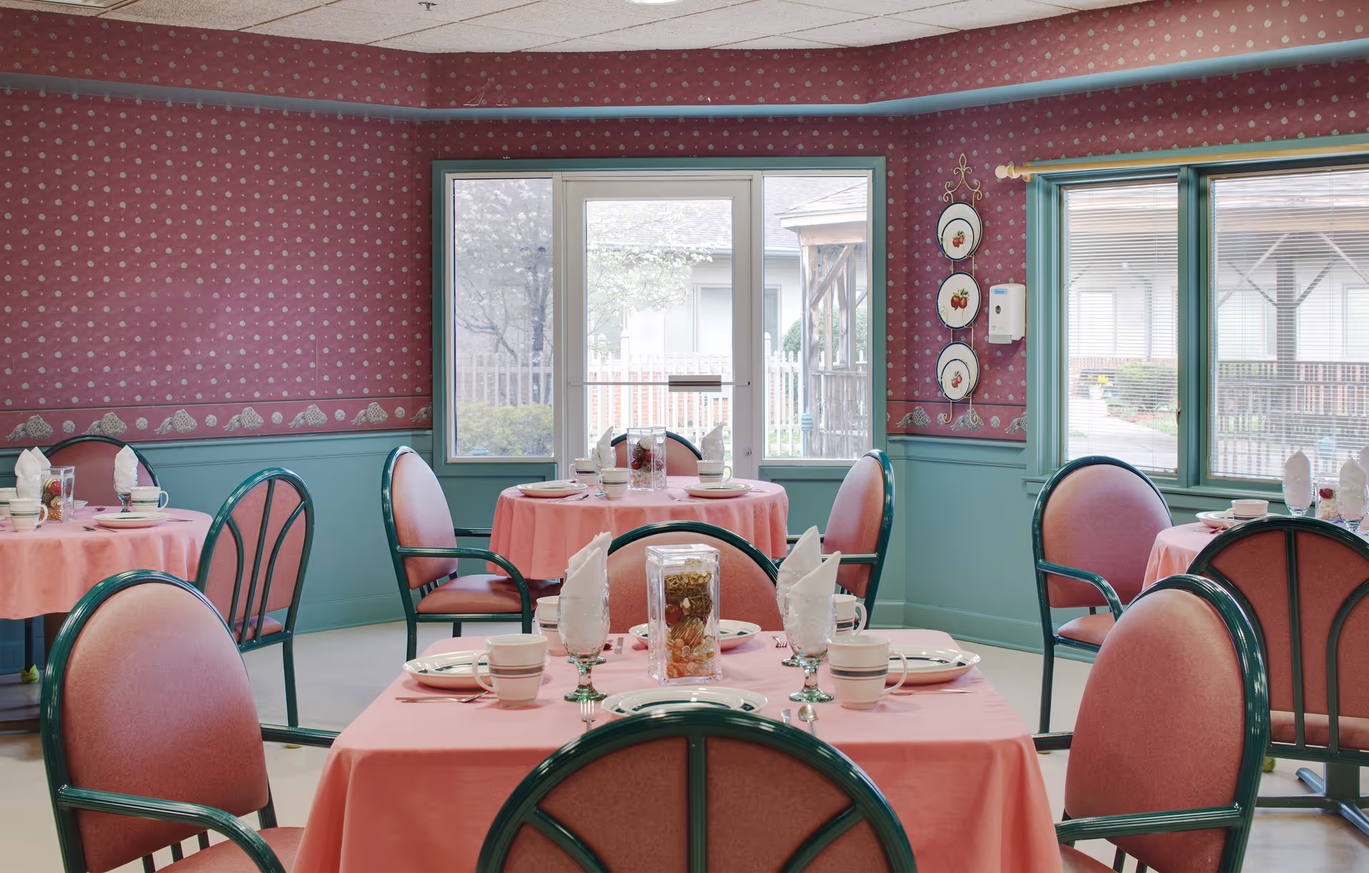 A dining room with several round tables covered in pink tablecloths, each set with plates, cups, and napkins. The chairs have green frames and pink cushions. The walls are decorated with red patterned wallpaper and teal wainscoting. Large windows allow natural light to enter the room.