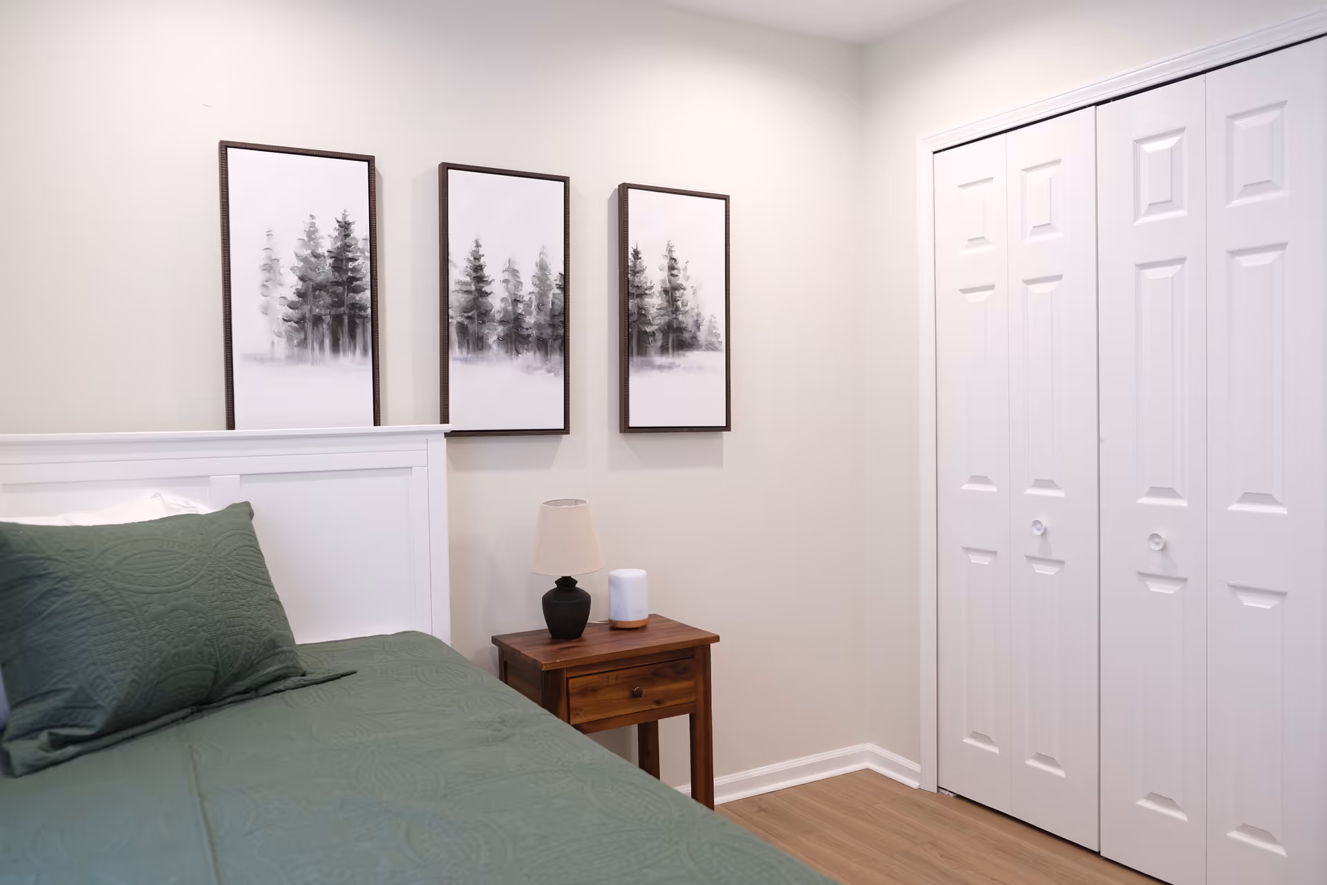 A clean bedroom with a white headboard, green bedding, a nightstand, and white closet doors.