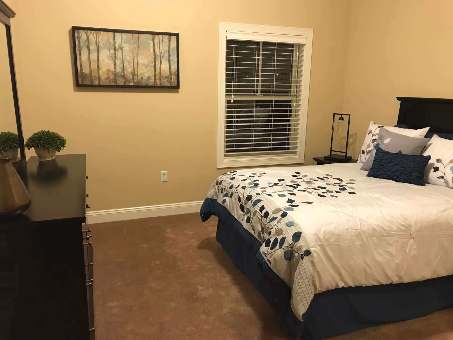 A bedroom with a bed covered in a white and blue floral comforter, several pillows, a window with white blinds, a nightstand with a lamp, a dresser with decorative items, and a framed painting of trees on the wall.