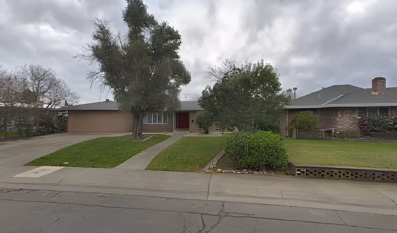 Single-story ranch-style house with a driveway, front lawn and trees under a cloudy sky.
