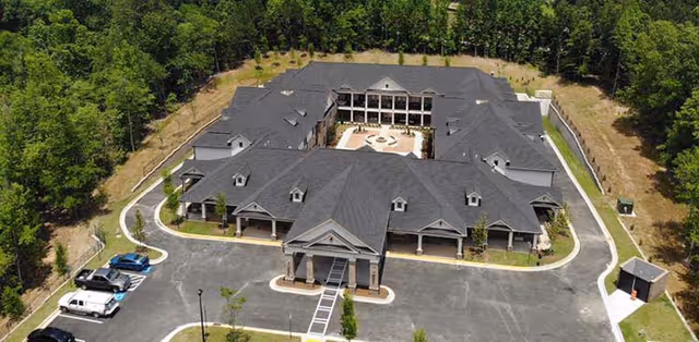 Aerial view of Magnolia Senior Living at Sugar Hill, showing a large U-shaped building with a central courtyard. The building is surrounded by trees and has a parking area with several vehicles. The roof is dark gray, and the exterior is light-colored with multiple windows and covered entrances.