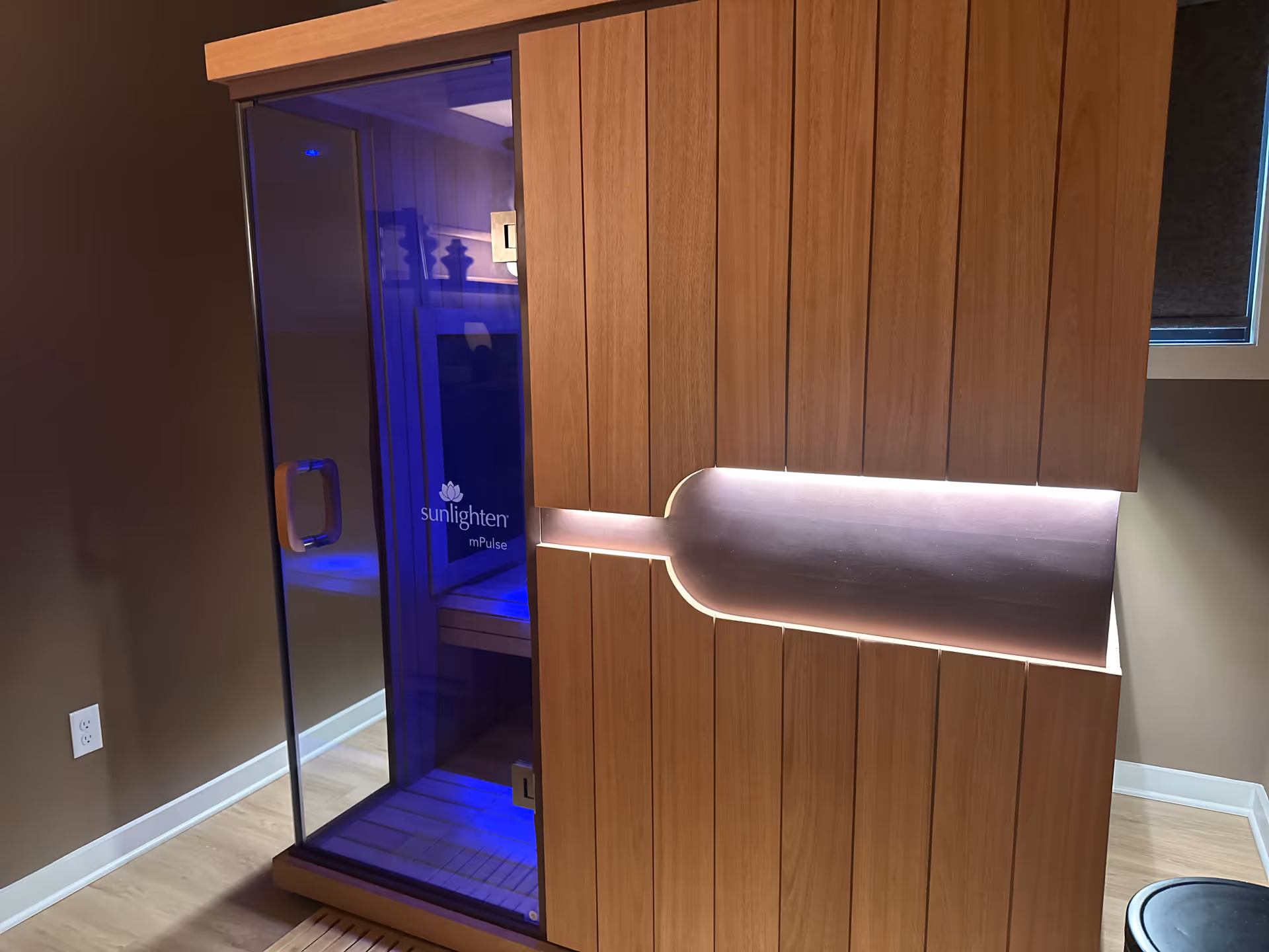 Wood-paneled interior with a glass Sunlighten sauna emitting blue light.