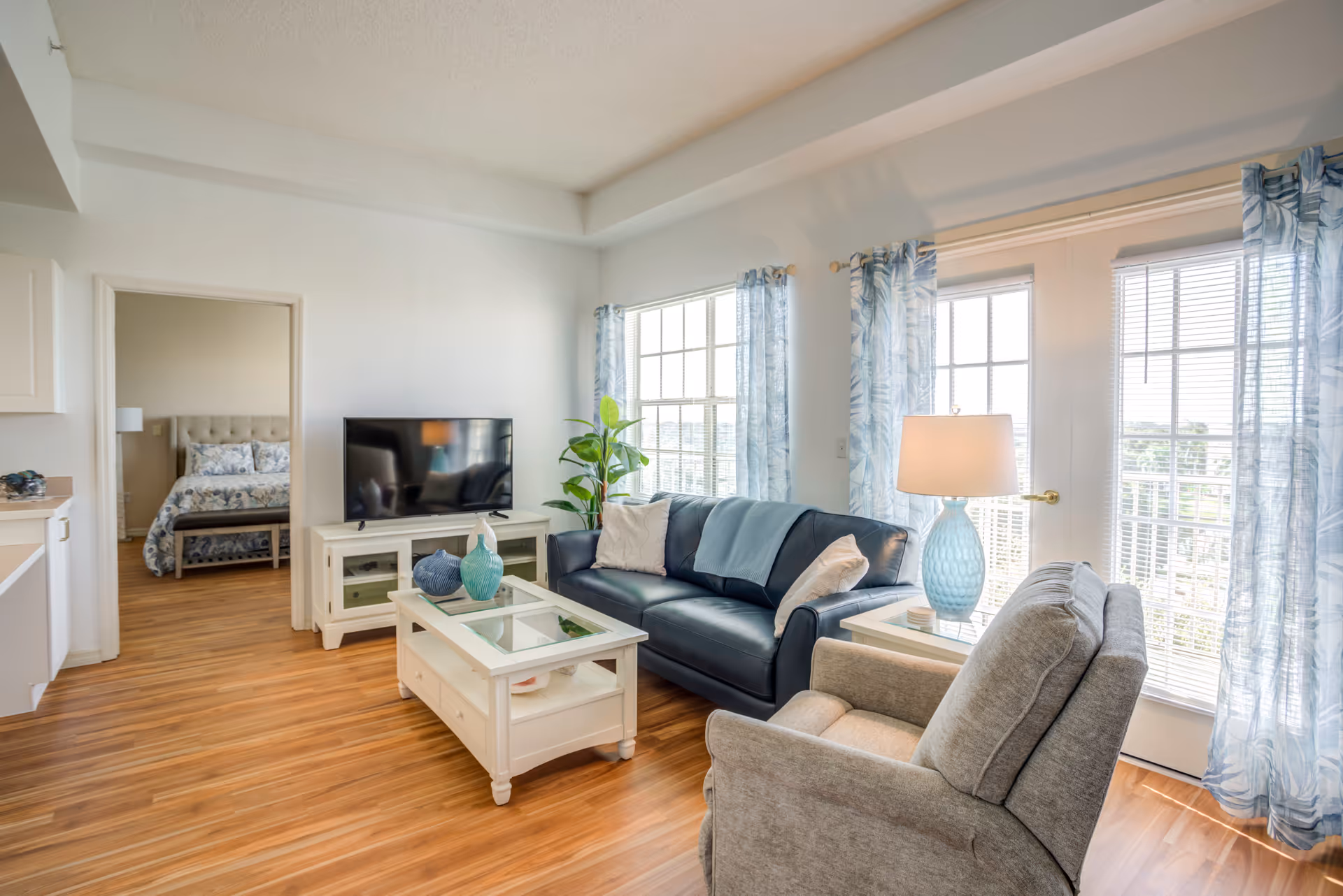 Bright living room in a senior facility with sofa, chairs, TV, and large windows.