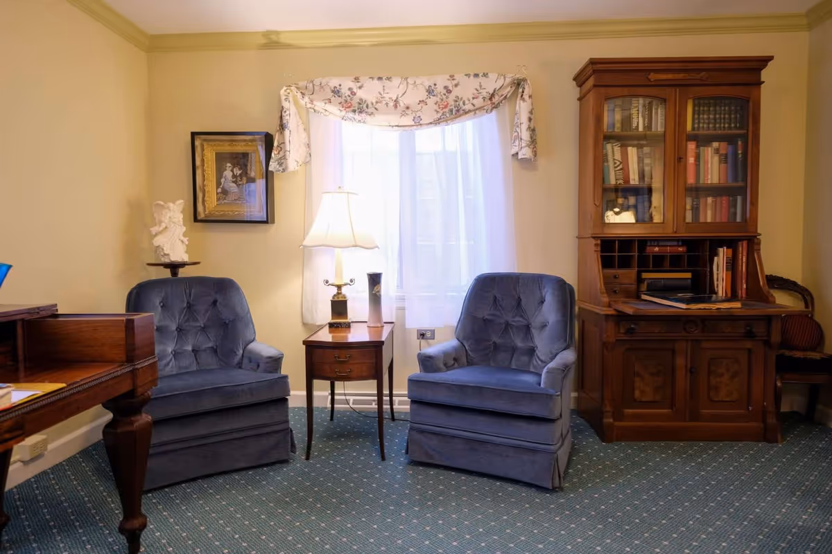 A cozy interior room with two blue upholstered armchairs placed on a patterned carpet. Between the chairs is a small wooden side table with a lamp and a decorative item. Behind the chairs is a window with sheer white curtains and a floral valance. To the right is a wooden cabinet with glass doors displaying books, and to the left is a wooden desk with carved legs. A framed picture and a small statue are on the left wall.