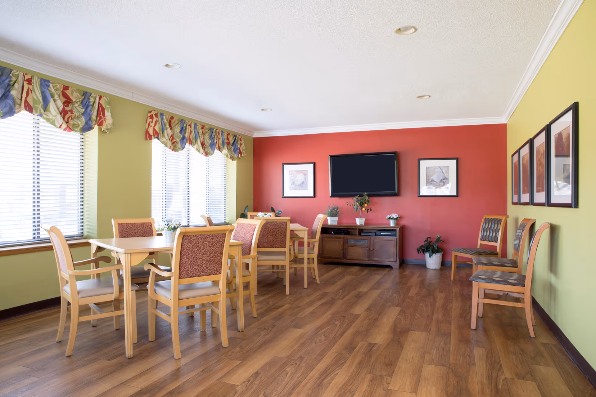 A bright and clean common area with wooden flooring, light green and red walls, and multiple chairs and tables arranged for seating. There are large windows with colorful valances allowing natural light to fill the room. A flat-screen TV is mounted on the red wall above a wooden cabinet, and framed artwork decorates the walls. Several potted plants add a touch of greenery to the space.