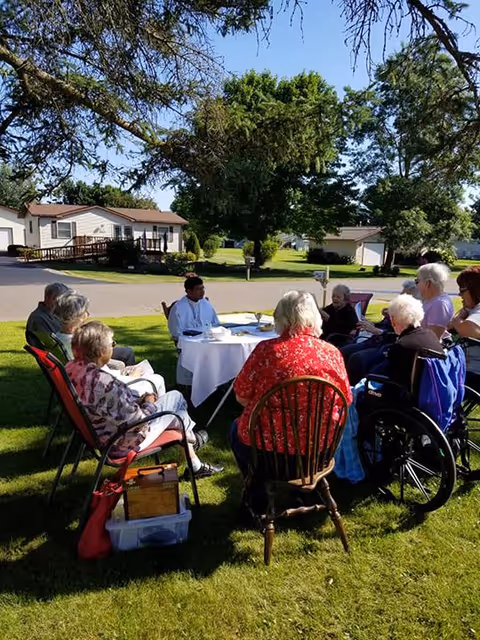 A group of elderly residents and a caregiver sit in a circle outdoors on a grassy lawn around a small table, with houses and trees in the background.