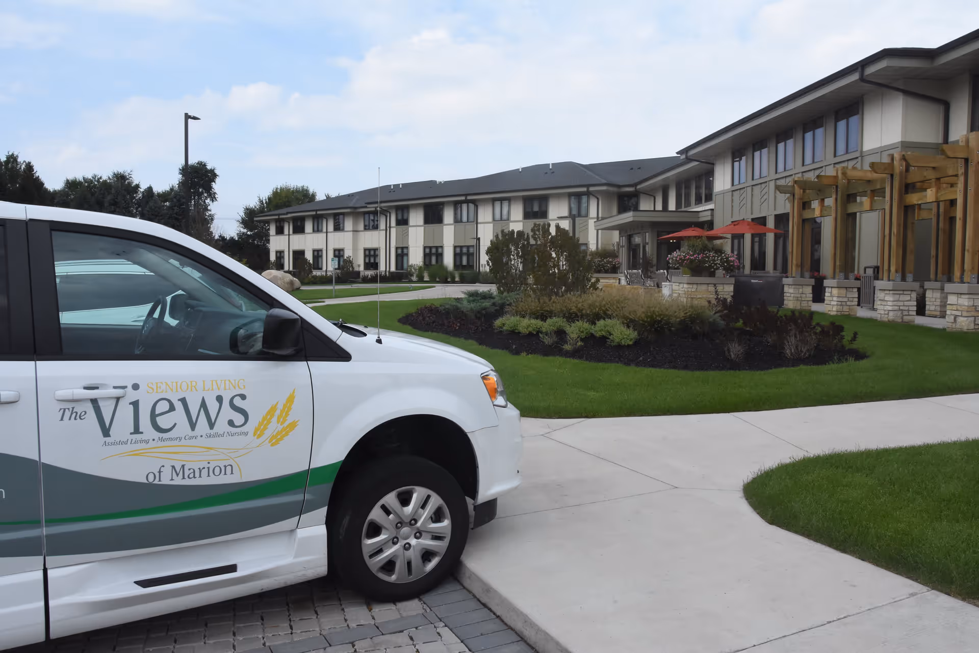 Exterior view of The Views Senior Living of Marion facility showing a white vehicle with the facility's logo parked on a paved area near a landscaped garden with green grass, shrubs, and a building with large windows and outdoor seating with red umbrellas in the background.