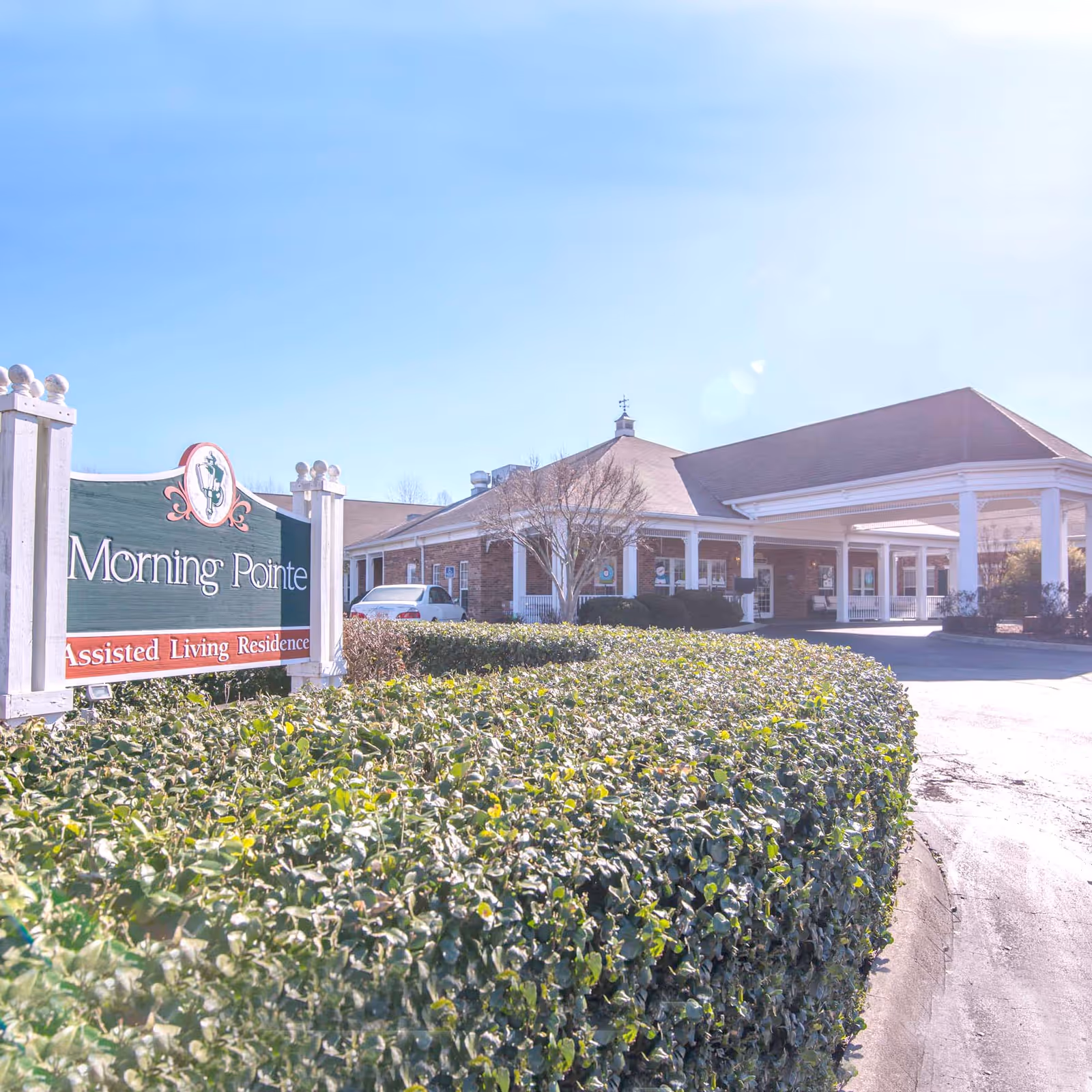Exterior view of Morning Pointe Assisted Living Residence building with a large covered entrance and a hedge in the foreground under a clear blue sky.