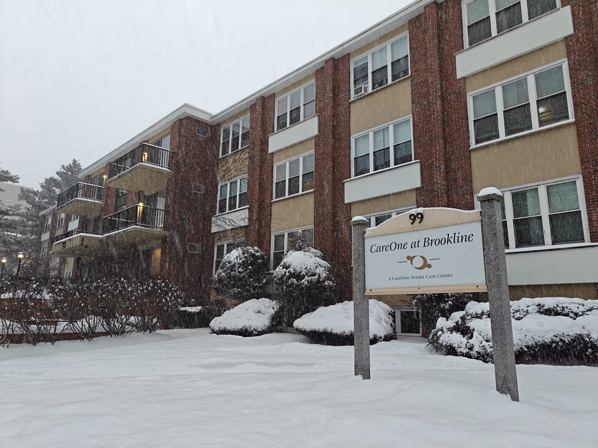Snow blankets the front exterior of CareOne at Brookline, a brick senior living building.