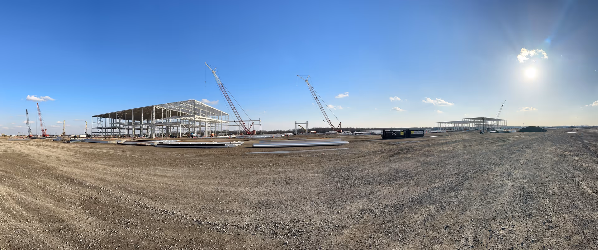 Construction site with steel framework and cranes on a dusty open lot under blue sky