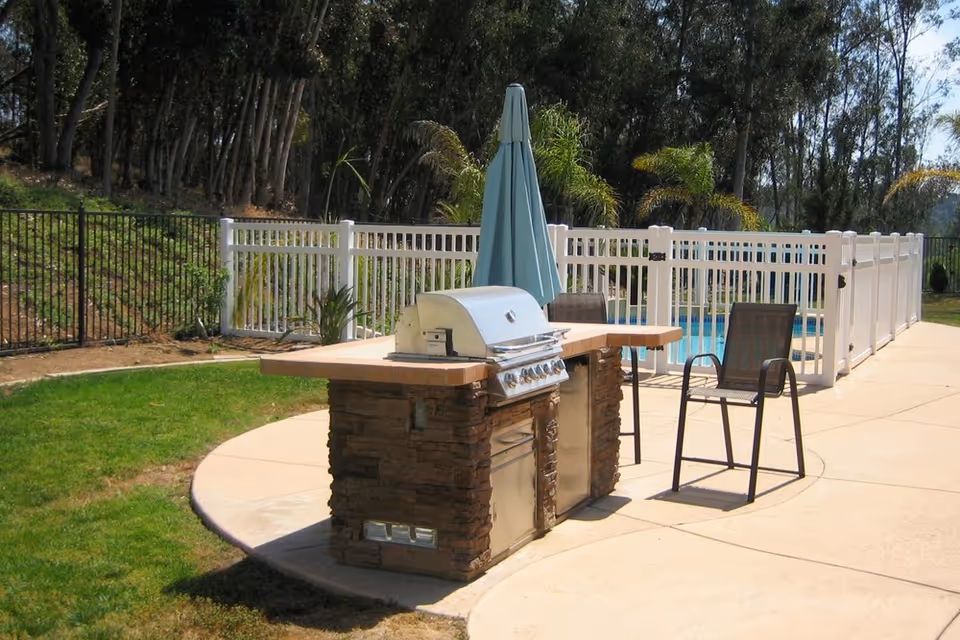 Outdoor patio area featuring a built-in stone and stainless steel barbecue grill with a closed blue umbrella and two metal chairs on a concrete surface. The area is enclosed by white fencing with a swimming pool visible behind it, and there are trees and greenery in the background.