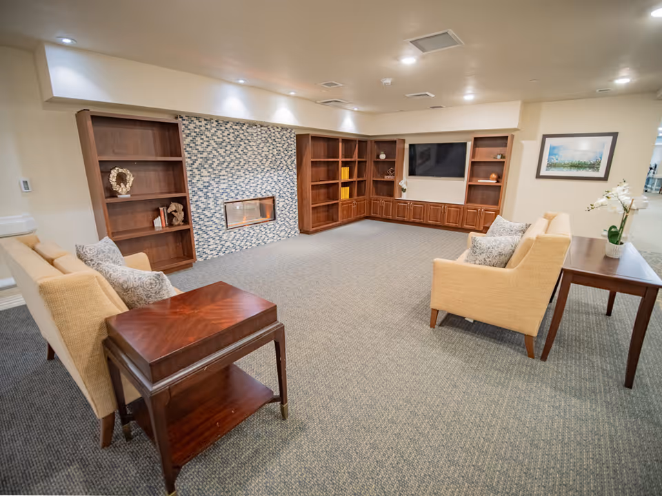 Cozy senior living lounge with beige seating, wooden shelves, and a fireplace.