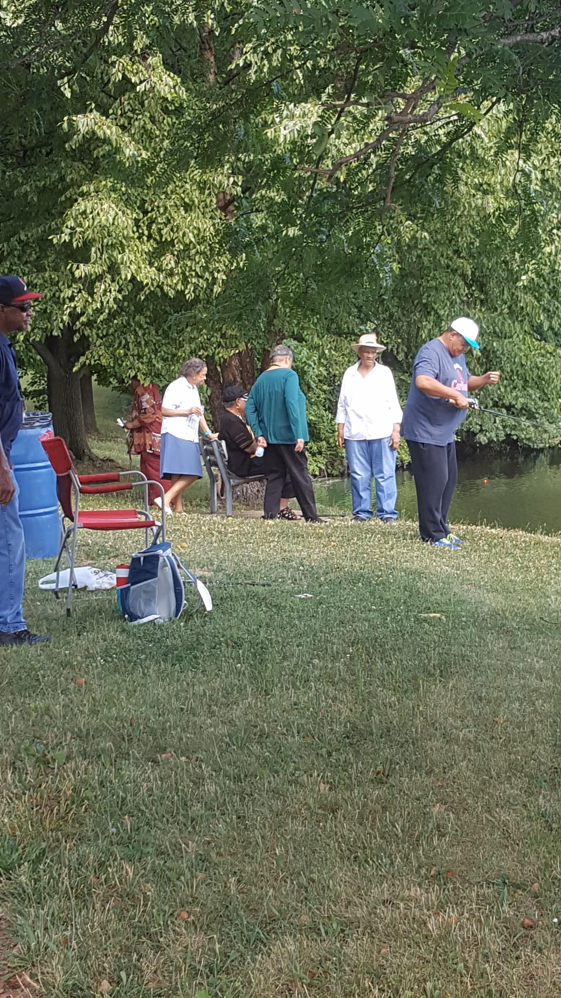 Group of seniors fishing and socializing near a pond under leafy trees