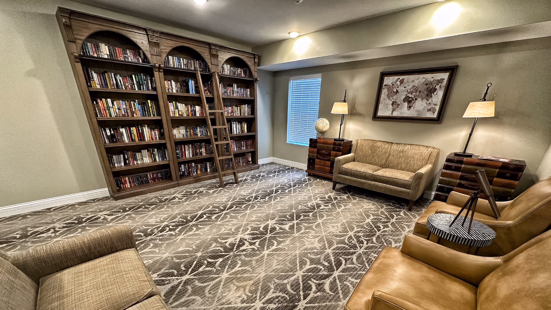 A cozy reading room with a large wooden bookshelf filled with books and a rolling ladder. The room has a patterned carpet, two beige sofas, two leather armchairs, two side tables with lamps, a globe, and a framed world map on the wall.