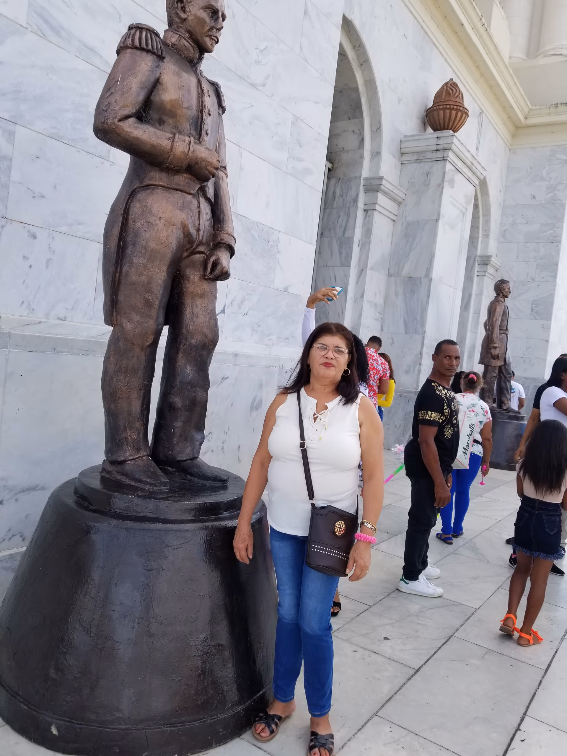 A woman stands beside a large statue outside a marble building with other visitors.
