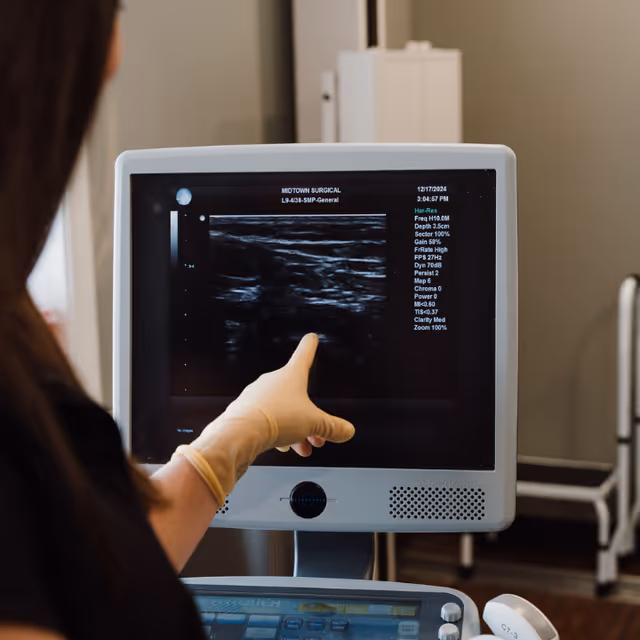 Gloved hand points to an ultrasound monitor in a clinical exam room.