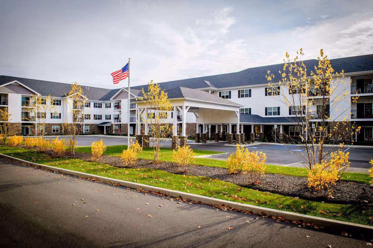 Front exterior of a multi-story senior living facility with a covered entrance, American flag, and autumn landscaping.