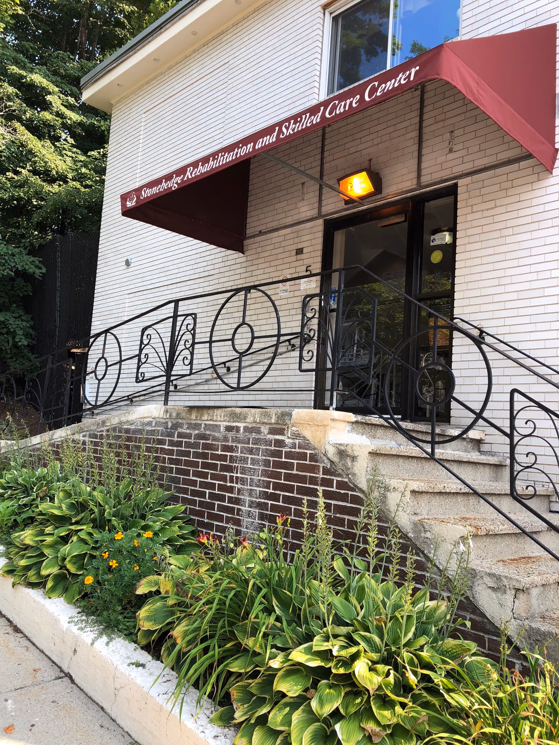 Entrance of Stonehedge Rehabilitation and Skilled Care Center with a maroon awning over glass doors, concrete steps, decorative iron railing, and a planted flowerbed.