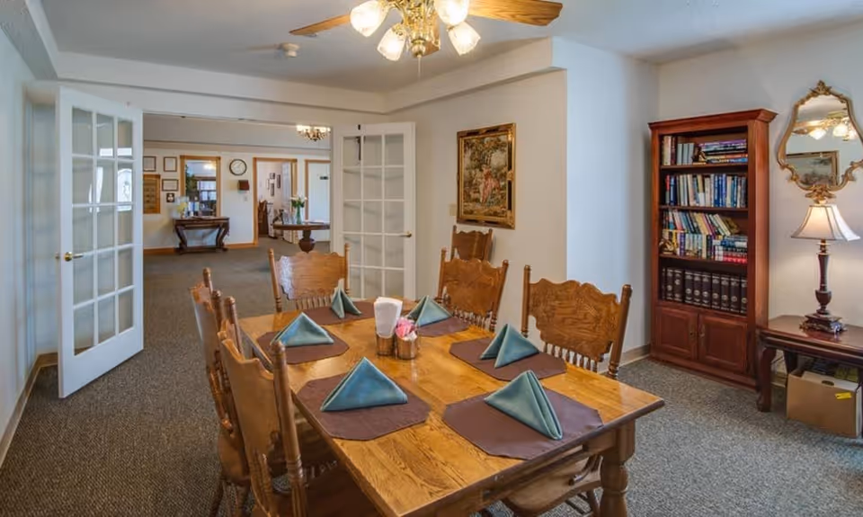 A dining area with a wooden table set for six people with folded blue napkins on brown placemats. The room has wooden chairs, a bookshelf filled with books, a table lamp, a mirror, and a framed painting on the wall. French doors open into another room with additional furniture and decor.
