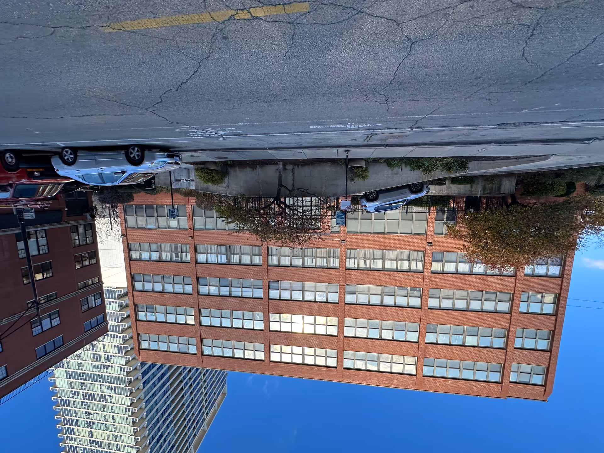 Exterior view of a multi-story brick building with many windows, a few trees, parked cars, and a clear blue sky.