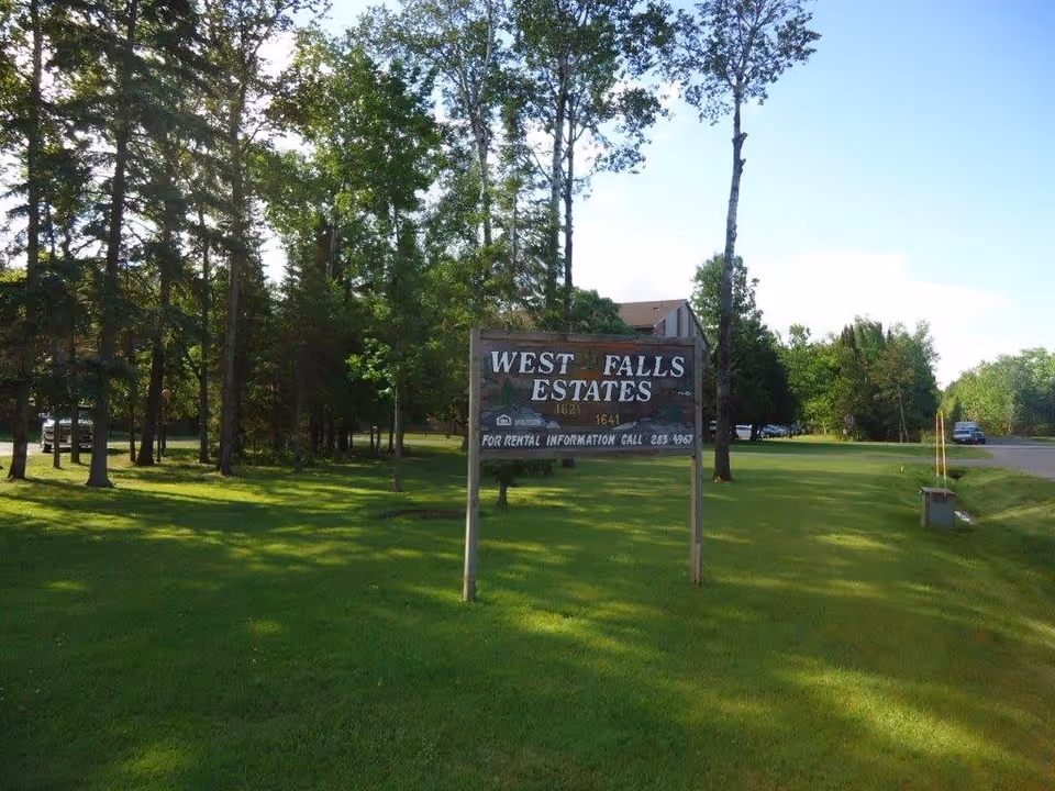 A wooden sign for West Falls Estates stands on a grassy area surrounded by trees, with a road and parked cars visible in the background under a clear sky.