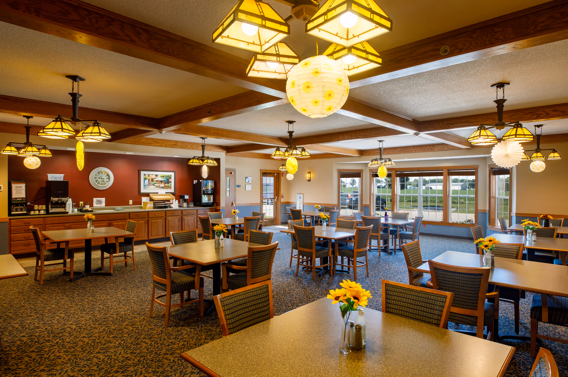 A bright and spacious dining room with multiple wooden tables and chairs arranged neatly. Each table has a small vase with yellow flowers. The room features large windows allowing natural light to enter, decorative ceiling lights, and a counter area with coffee machines and other amenities against a red accent wall.