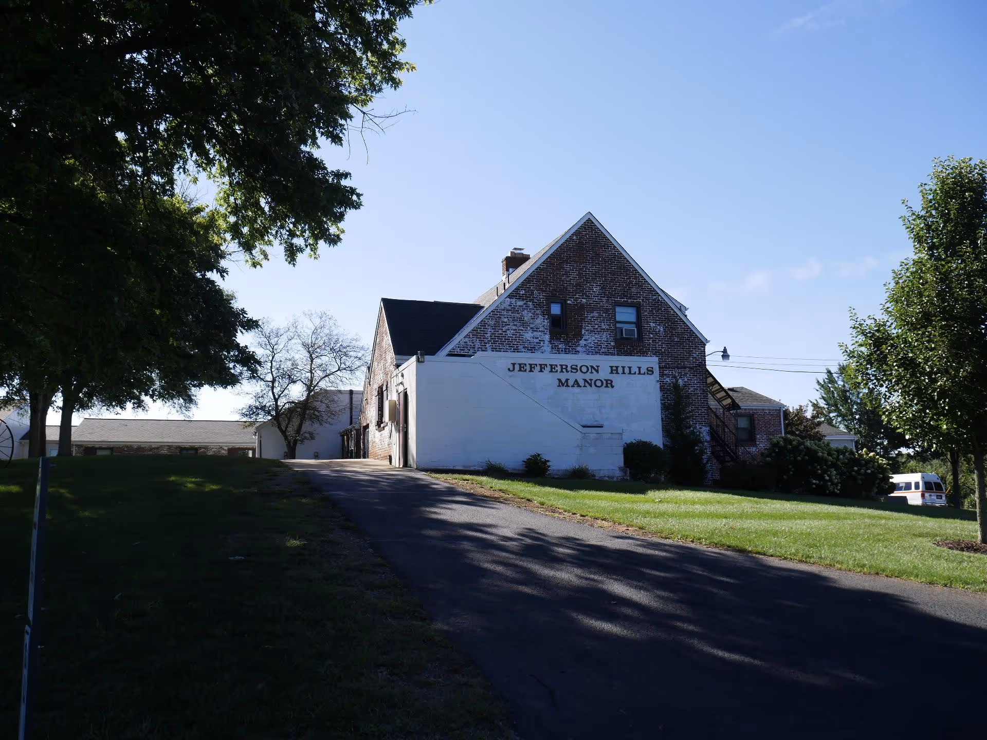 Brick and white building labeled "Jefferson Hills Manor" at the end of a driveway with trees and lawn under a clear sky.