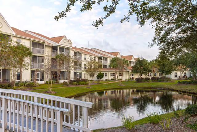 Exterior view of a multi-story senior living facility named Brookdale Destin with balconies, surrounded by landscaped greenery and a small pond with a white wooden bridge in the foreground.