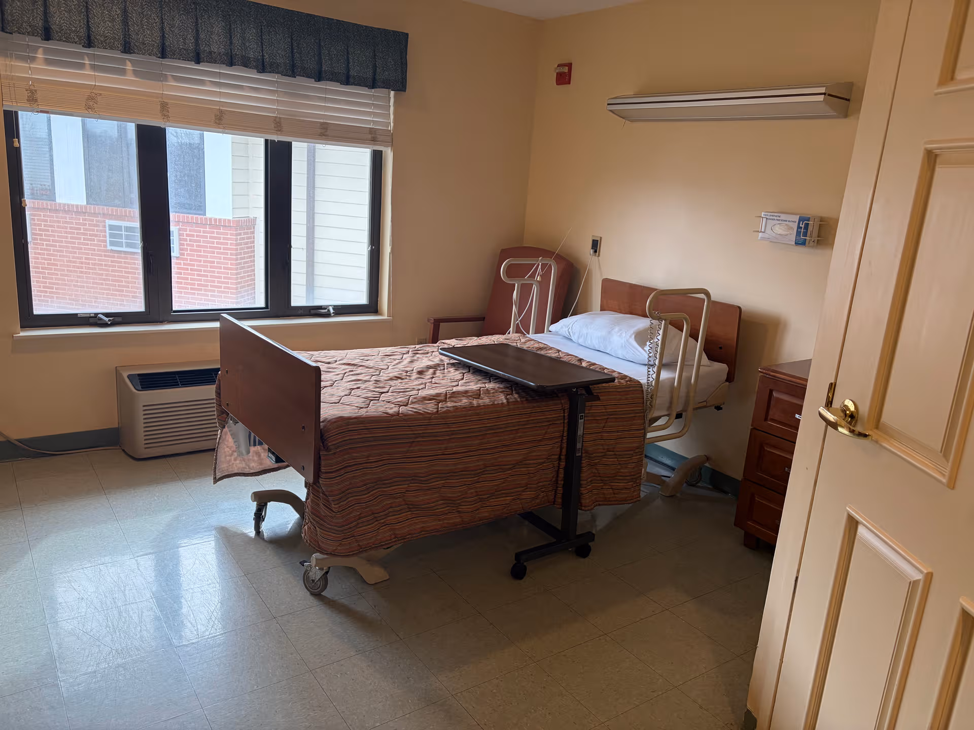 A patient bedroom in a senior facility with bed, window, and furniture.