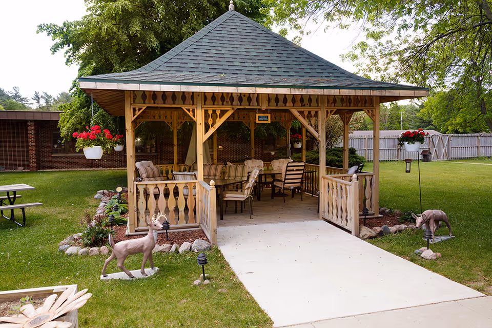 Wooden gazebo with chairs and hanging flower baskets on a grassy lawn in front of a brick building.