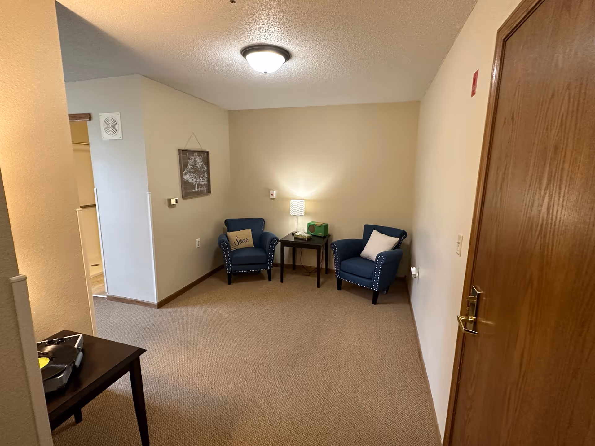 A small sitting area with two blue armchairs, each with a decorative pillow, positioned around a small dark wooden side table with a lamp and a green radio. The room has beige walls and carpet, a wooden door on the right, and a framed picture hanging on the left wall. There is a ceiling light fixture and an open doorway leading to another room.