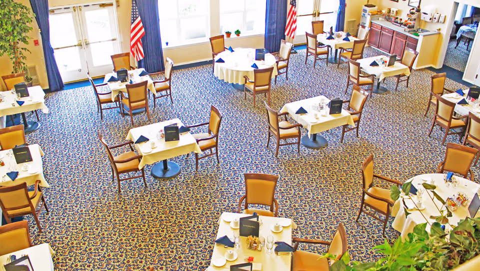 A bright dining room with multiple set tables and chairs on patterned carpet and American flags by the windows.