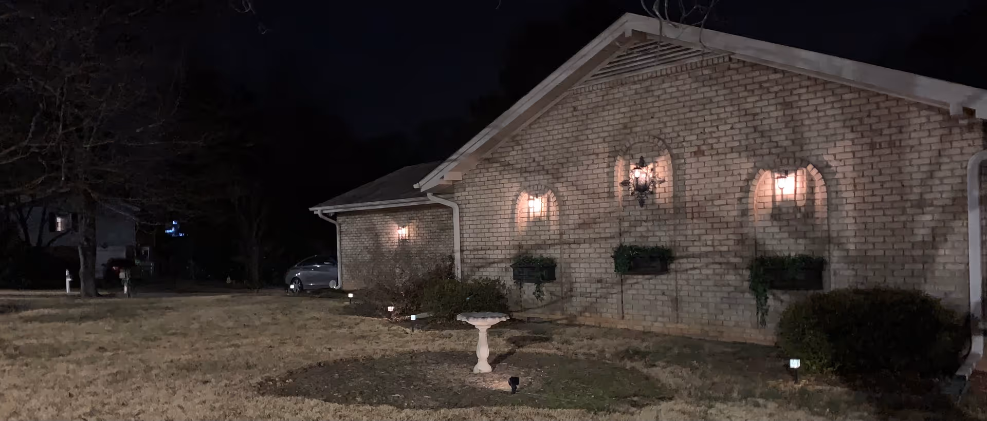 Night view of the side exterior of a brick building with three arched windows, each with a light above and planter boxes below. There is a small birdbath in the grass in front of the building, and a car is parked near the corner. The surrounding area includes some bushes and leafless trees.