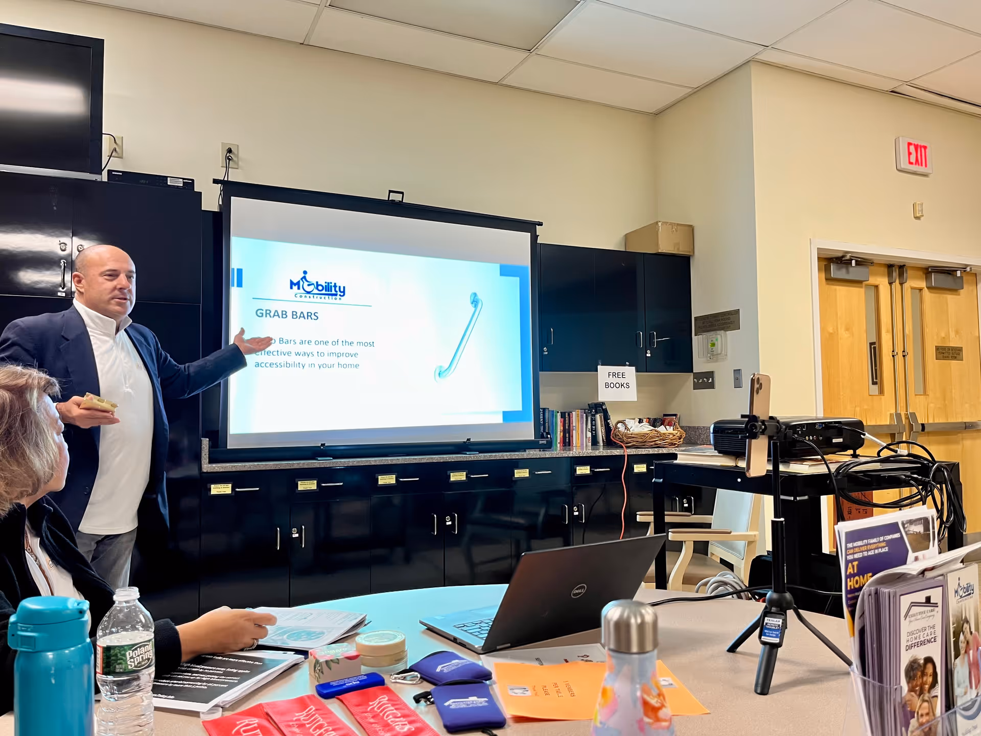 Presenter in a suit lectures beside Mobility & Motion LLC slide on projector in conference room.