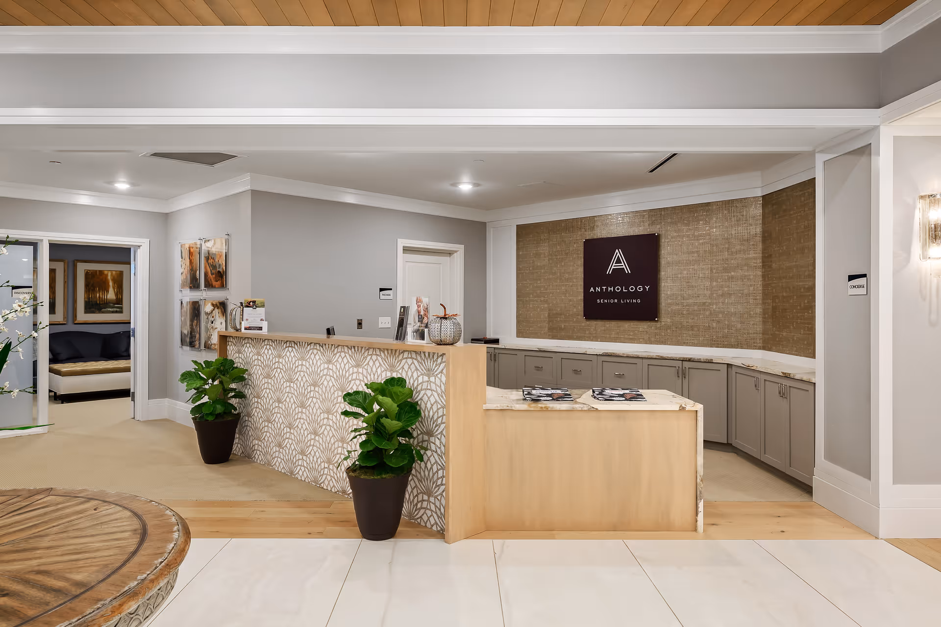 Reception area of a senior living facility with a wooden front desk featuring a patterned front panel, two potted plants on the floor, and a sign on the back wall that reads 'Anthology Senior Living'. The area has light-colored walls, recessed lighting, and a glimpse into an adjacent room with artwork and seating.