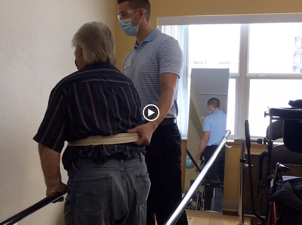 A healthcare worker wearing a mask and protective glasses assists an elderly man with a gait belt as he uses parallel bars for walking practice in a rehabilitation setting. There is a large window and a mirror in the background reflecting the scene.