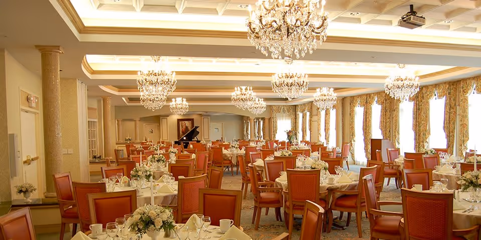 A spacious, elegant dining room with multiple round tables covered in white tablecloths, each set with plates, glasses, napkins, and floral centerpieces. The room features large windows with patterned curtains, ornate chandeliers hanging from the ceiling, and a grand piano in the background.
