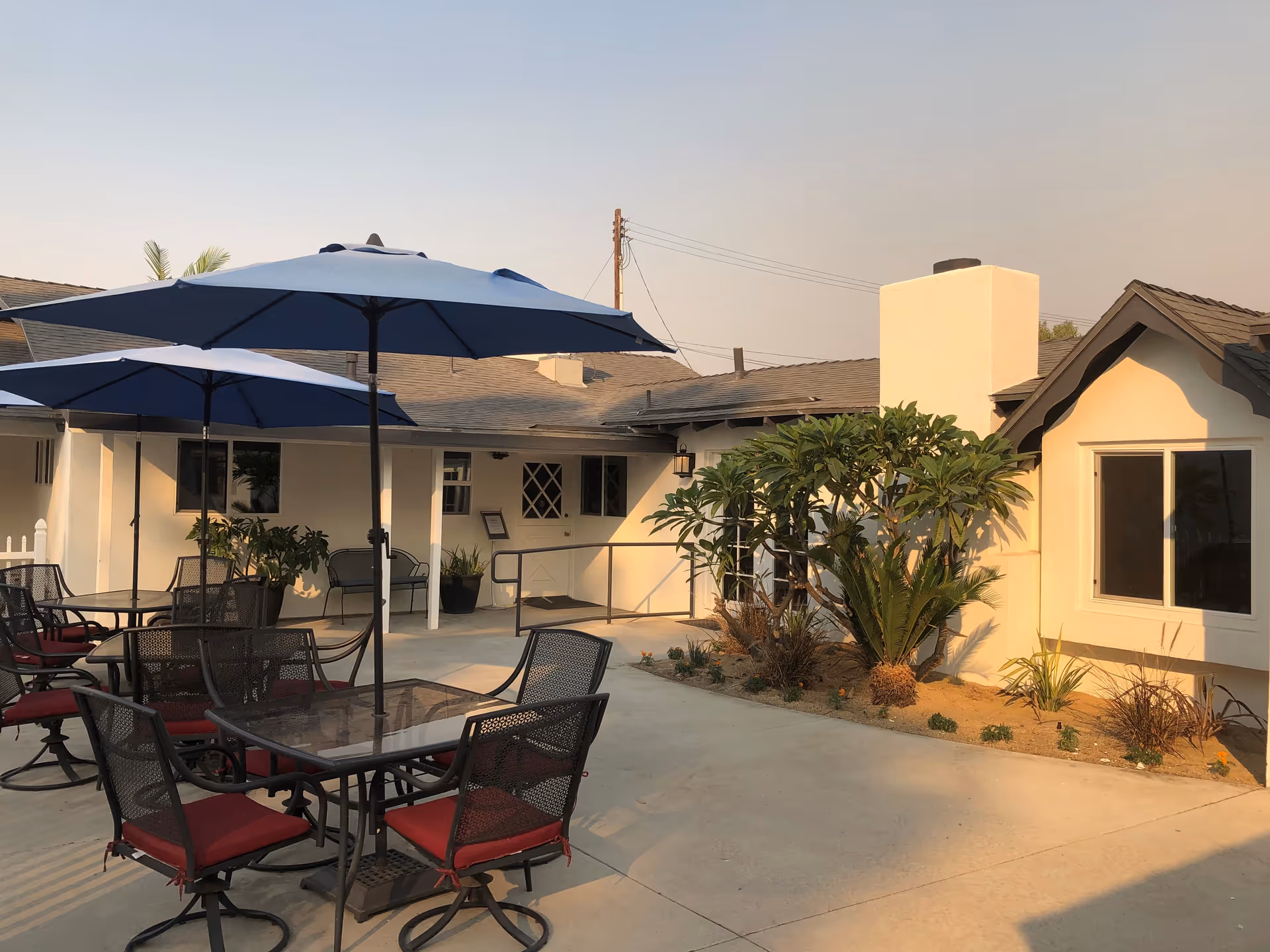 Outdoor courtyard with tables, chairs, blue umbrellas, and surrounding single-story building.