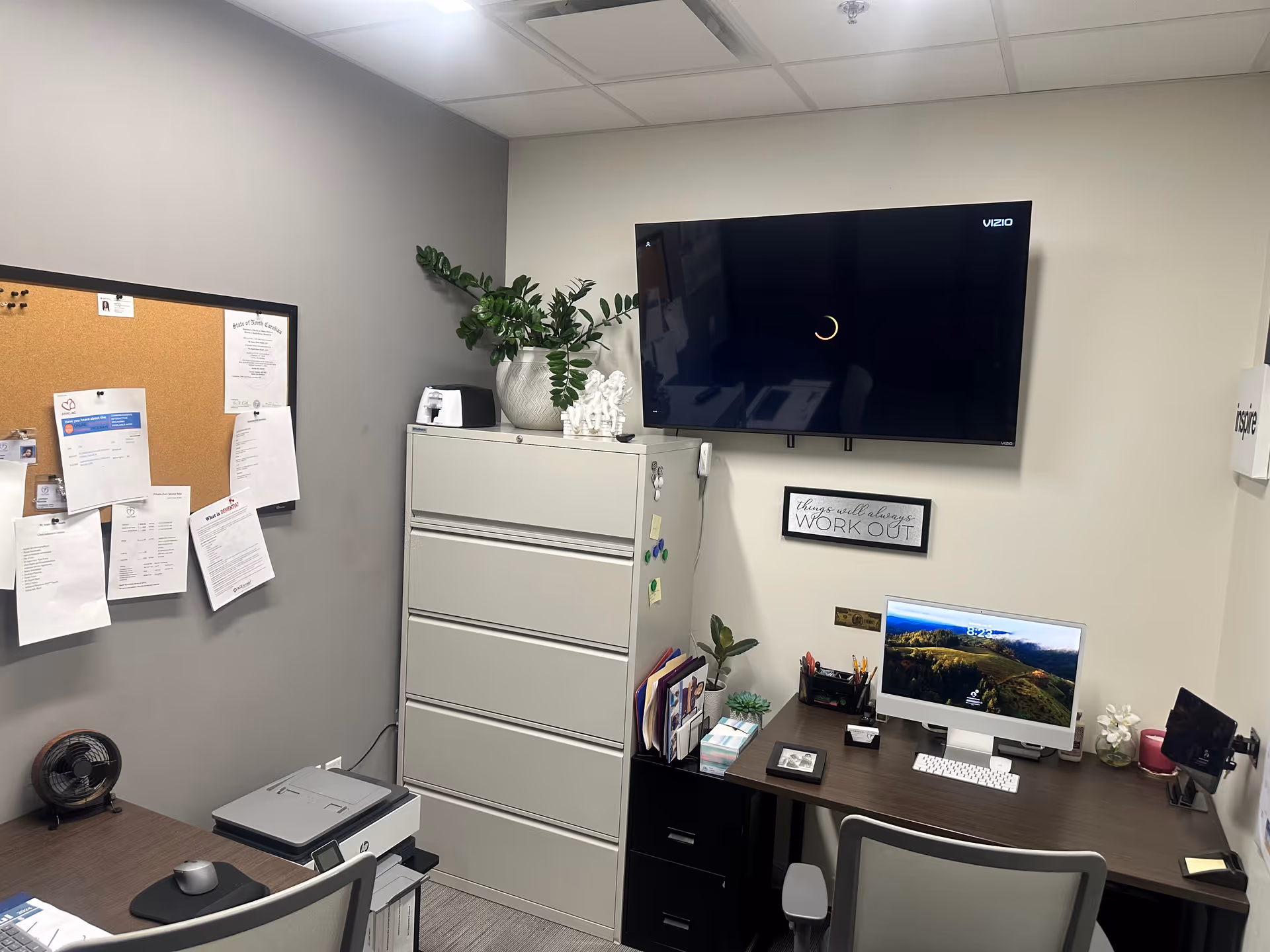 Small office workspace with desk, computer, filing cabinet, corkboard, plant, and wall-mounted TV.