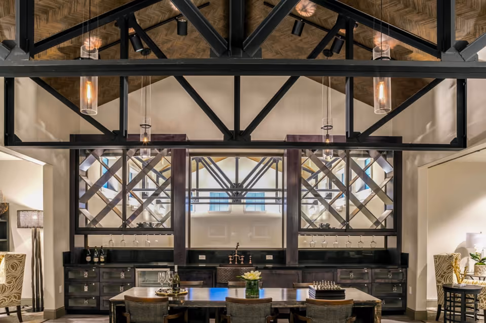 Communal dining area with a long wooden table and chairs in front of a built-in bar and wine racks beneath exposed vaulted ceiling beams.