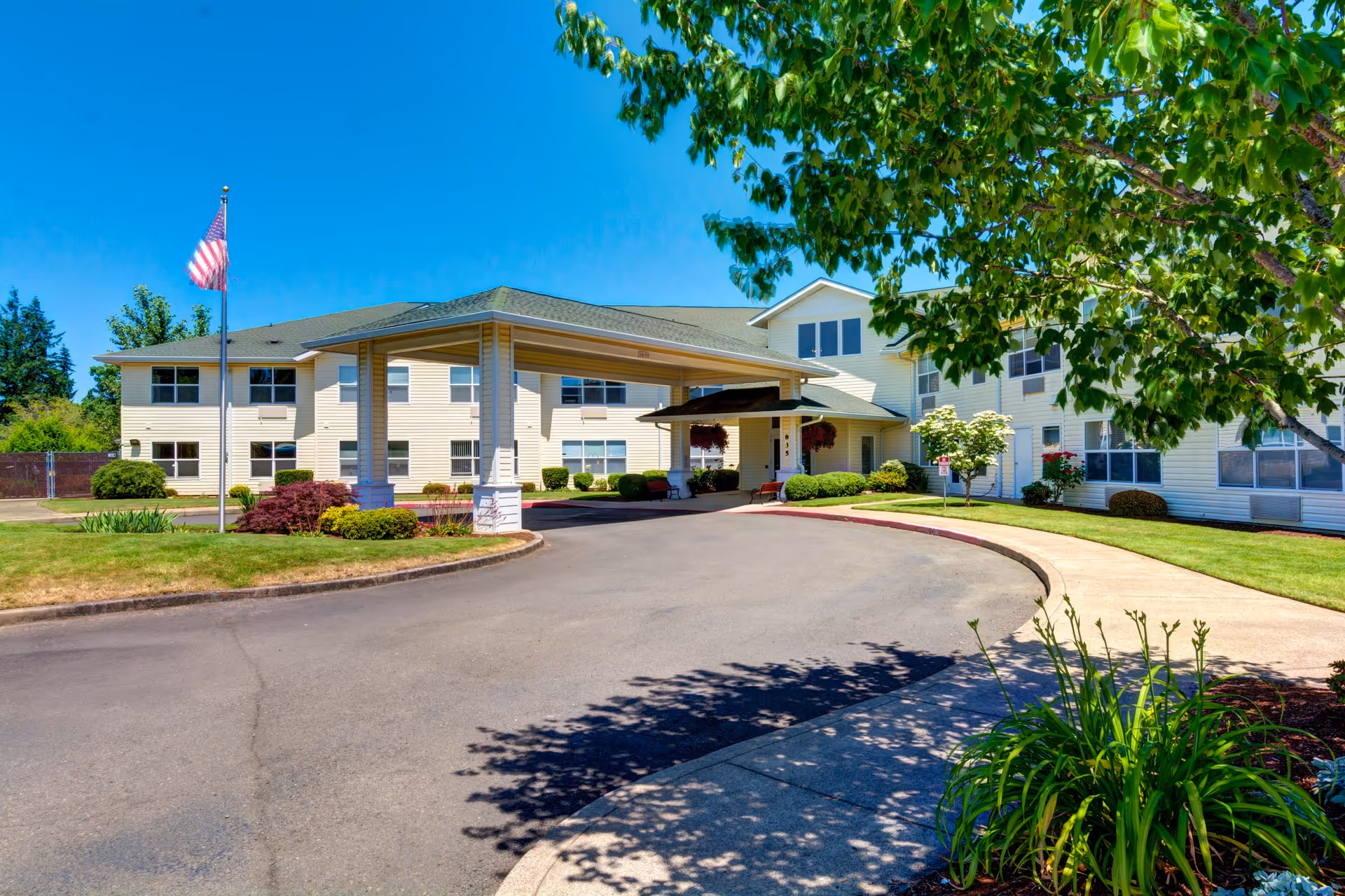 Exterior view of a two-story senior living facility building with a covered entrance driveway, an American flag on a flagpole, well-maintained landscaping including bushes, trees, and grass, under a clear blue sky.