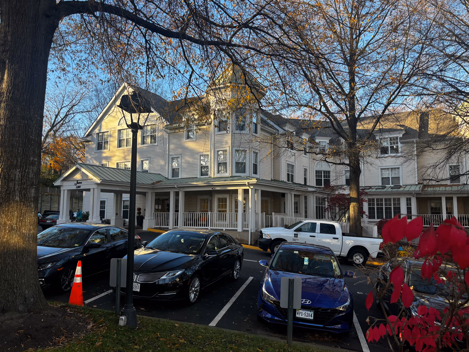 Exterior view of a multi-story senior living facility building with white siding and a green metal roof. Several cars are parked in front of the building, and there are leafless trees and some red foliage visible. The building has a covered entrance with white columns and a porch area.