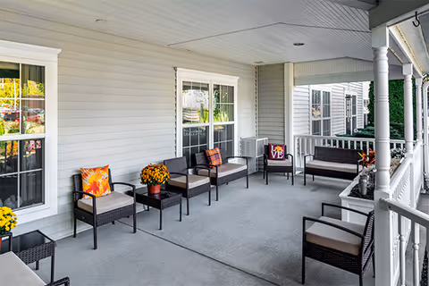 Covered outdoor porch area with multiple cushioned wicker chairs and small tables, decorated with colorful pillows and potted flowers, attached to a light gray building with white trim and windows.