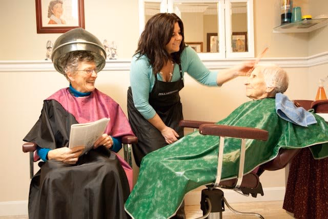 A hair salon scene in an assisted living center where a stylist is brushing the hair of an elderly woman seated in a salon chair covered with a green cape, while another elderly woman sits nearby under a hair dryer with a pink cape, holding a newspaper and smiling.